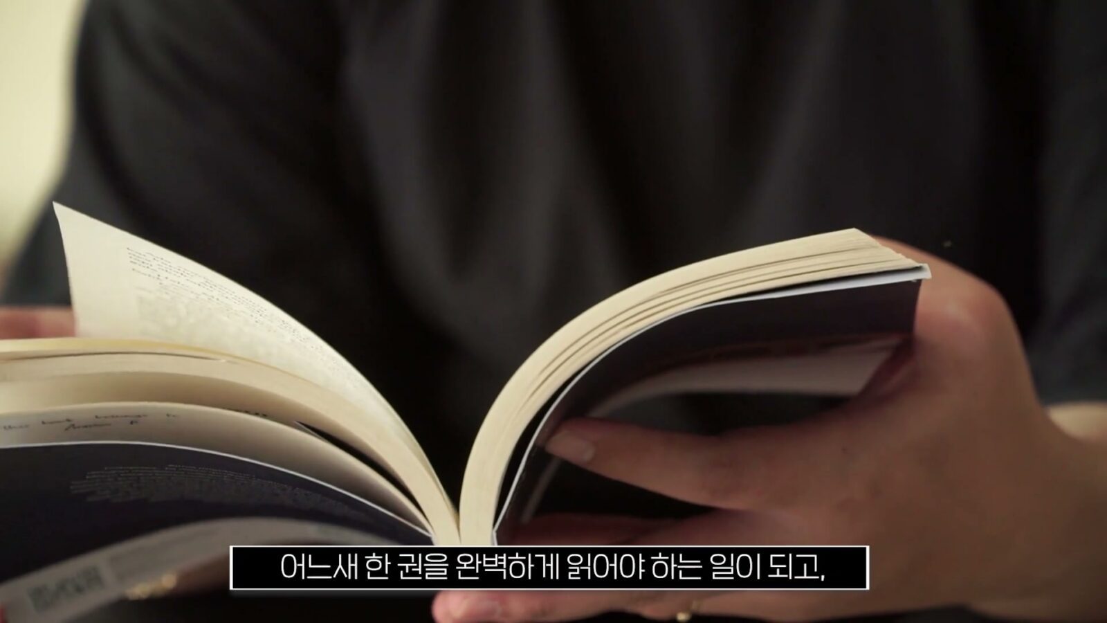 어두운 옷을 입은 사람이 펼친 책의 페이지를 손으로 넘기고 있고, 하단 자막 바에 '어느새 한 권을 완벽하게 읽어야 하는 일이 되고,'라고 적힌 화면